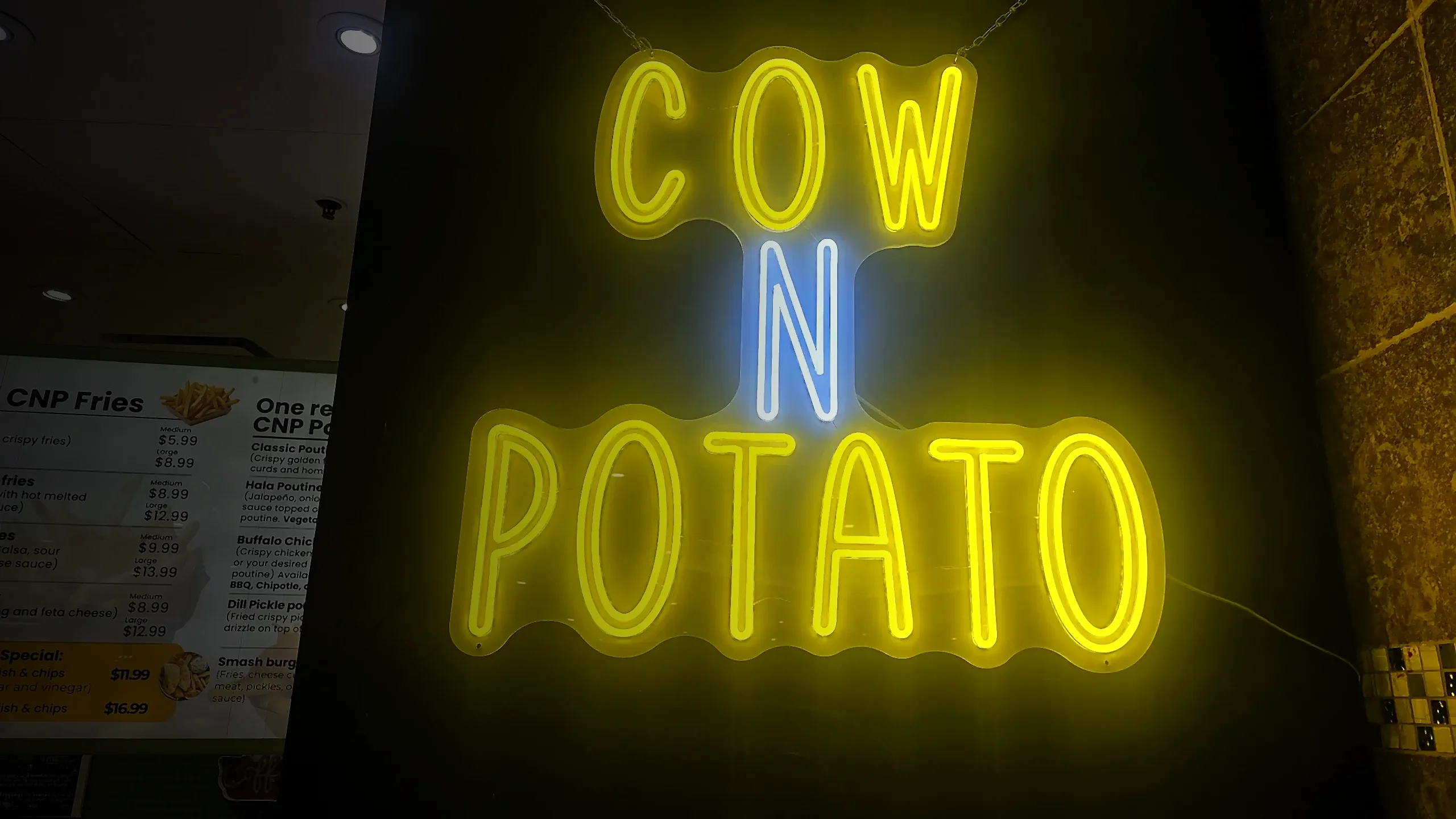 Yellow and blue COW N POTATO neon sign glowing on a black wall beside the menu board